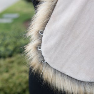 May include: A beige faux fur vest with a brown fur trim. The vest has four silver metal rings on the side.