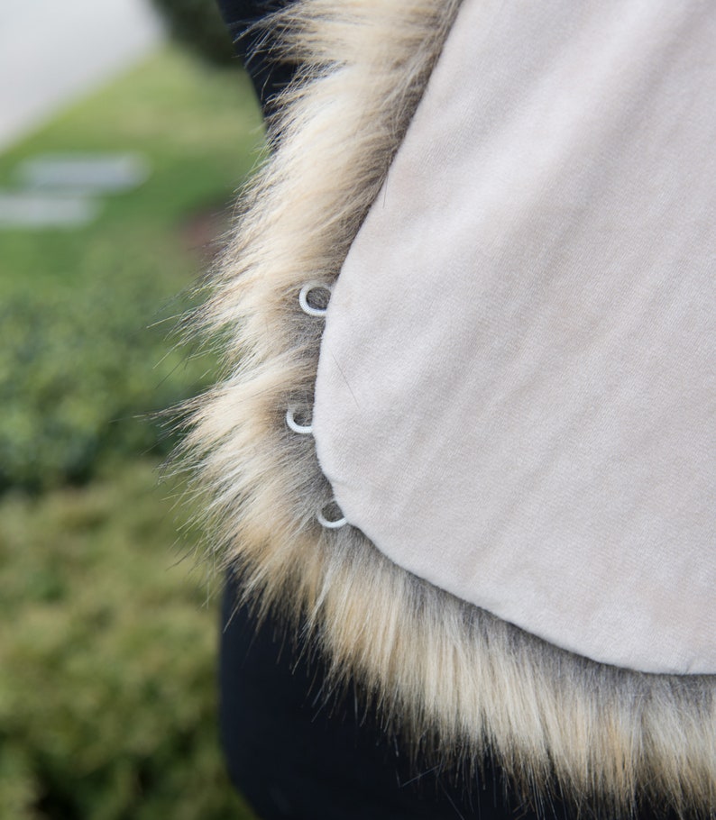 May include: A beige faux fur vest with a brown fur trim. The vest has four silver metal rings on the side.