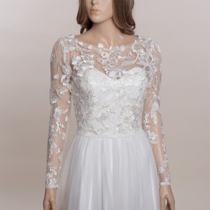 Op de afbeelding: Een witte trouwjurk met een sweetheart halslijn en een lange mouwen kanten overlay. De overlay heeft bloemenborduurwerk en doorschijnende mouwen. De rok is gemaakt van een vloeiende, gelaagde stof.