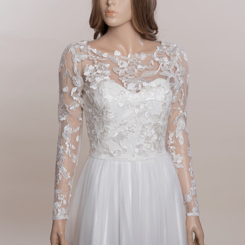 Op de afbeelding: Een witte trouwjurk met een sweetheart halslijn en een lange mouwen kanten overlay. De overlay heeft bloemenborduurwerk en doorschijnende mouwen. De rok is gemaakt van een vloeiende, gelaagde stof.