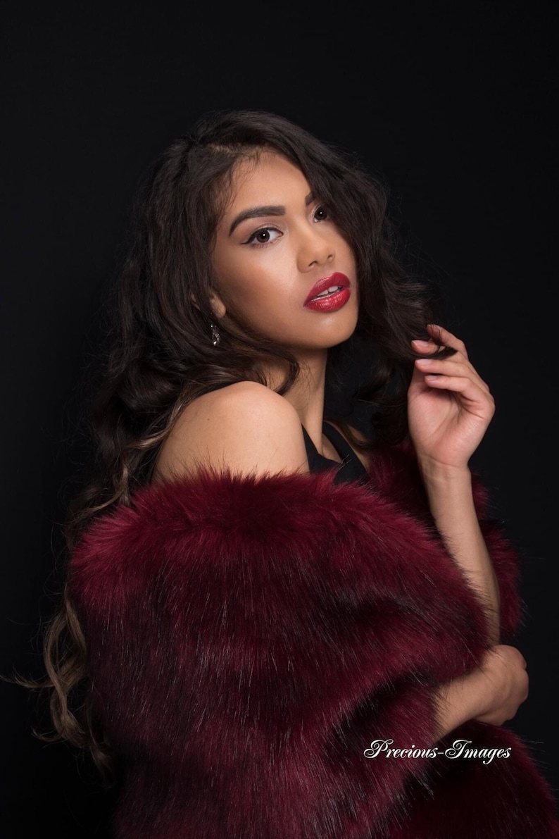 May include: A woman with long dark hair wearing a burgundy faux fur shawl. She is looking over her shoulder with a slight smile and wearing red lipstick.
