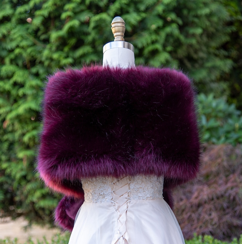 May include: A deep purple faux fur shawl with a soft, fluffy texture. The shawl is draped over a white dress with a lace bodice.