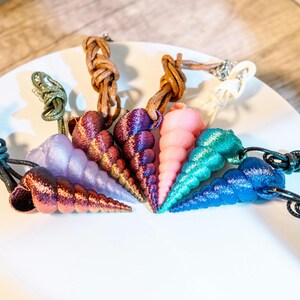 May include: A collection of colorful, cone-shaped pendants, resembling seashells, arranged on a white plate. The pendants are in shades of purple, pink, blue, and green, each with a textured surface. They are attached to cords of various colors.