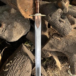 May include: A detailed sword with a silver blade and a bronze hilt. The hilt features a decorative design and a textured grip. The sword is displayed against a backdrop of wooden logs, showcasing its craftsmanship.