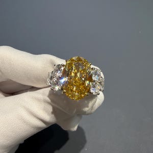 Puede incluir: Un lujoso anillo con un gran diamante amarillo de talla ovalada como pieza central, flanqueado por dos diamantes claros m&aacute;s peque&ntilde;os a cada lado. El anillo est&aacute; engastado en una banda de oro, mostrando el brillo de las piedras preciosas.