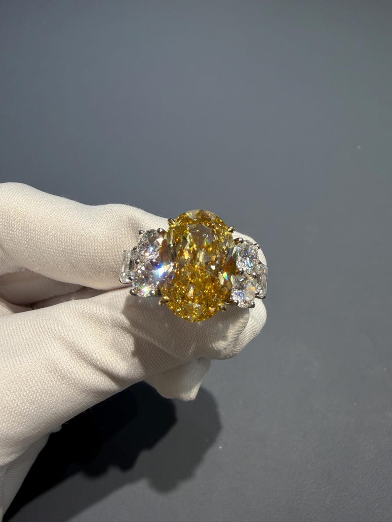 Puede incluir: Un lujoso anillo con un gran diamante amarillo de talla ovalada como pieza central, flanqueado por dos diamantes claros m&aacute;s peque&ntilde;os a cada lado. El anillo est&aacute; engastado en una banda de oro, mostrando el brillo de las piedras preciosas.