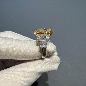 Puede incluir: Un anillo de platino y oro con un gran diamante amarillo de talla ovalada. El anillo tiene un halo de diamantes m&aacute;s peque&ntilde;os y una banda con acentos de diamantes adicionales. El anillo es sostenido por una mano enguantada sobre un fondo gris.