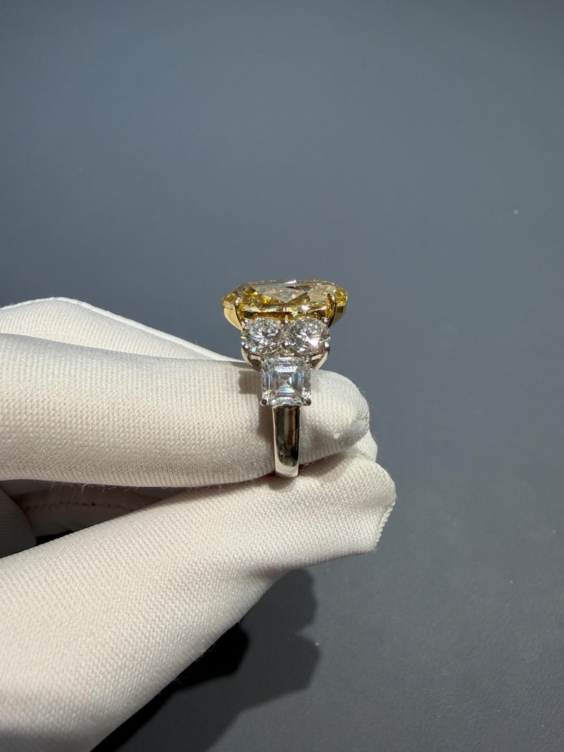 Puede incluir: Un anillo de platino y oro con un gran diamante amarillo de talla ovalada. El anillo tiene un halo de diamantes m&aacute;s peque&ntilde;os y una banda con acentos de diamantes adicionales. El anillo es sostenido por una mano enguantada sobre un fondo gris.