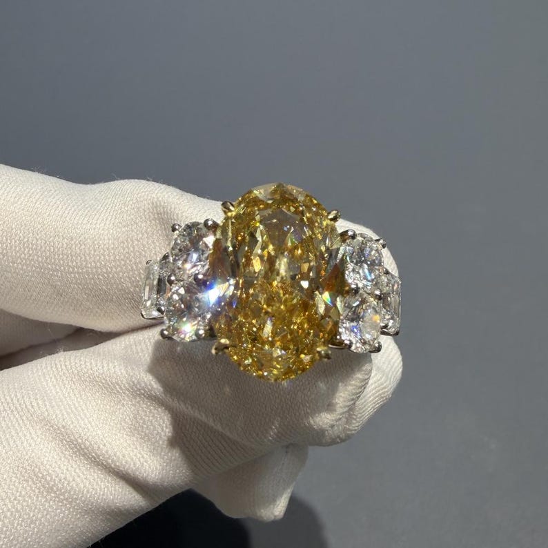 Puede incluir: Un lujoso anillo con un gran diamante amarillo de talla ovalada como pieza central, flanqueado por grupos de diamantes m&aacute;s peque&ntilde;os y claros. El anillo est&aacute; engarzado en un metal blanco, probablemente platino u oro blanco, y se muestra sobre un fondo neutro.