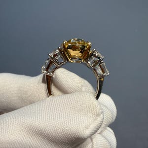 Puede incluir: Primer plano de un anillo con una gran piedra preciosa amarilla redonda como pieza central. El anillo tiene una banda plateada con piedras preciosas rectangulares y transparentes a cada lado de la piedra amarilla. El anillo es sostenido por una mano enguantada sobre un fondo gris.