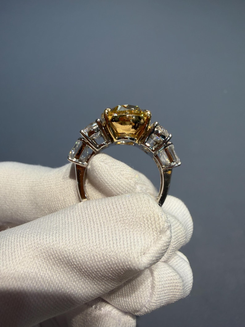 Puede incluir: Primer plano de un anillo con una gran piedra preciosa amarilla redonda como pieza central. El anillo tiene una banda plateada con piedras preciosas rectangulares y transparentes a cada lado de la piedra amarilla. El anillo es sostenido por una mano enguantada sobre un fondo gris.