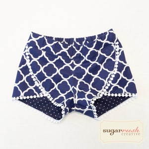 May include: Navy blue shorts featuring a white quatrefoil pattern and an elastic waistband. The leg openings are accented with white pom-pom trim and reveal a dark blue fabric with white polka dots. The text sugar rush creative is visible.