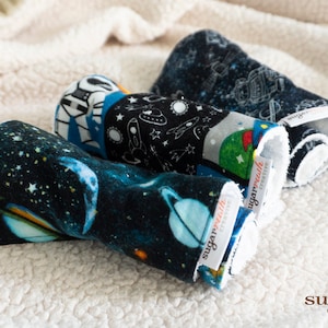 May include: Three rolled up burp cloths with a space theme. The cloths are black with blue and white stars, planets, and rockets. The cloths have a white label with the text "sugar rush creative" on them.