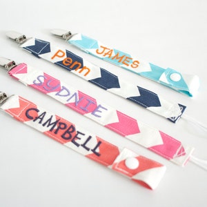 May include: Four personalized pacifier clips with white and colored chevron patterns. The clips are labeled "James", "Penn", "Sydnie", and "Campbell".