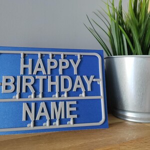 May include: A blue card with white plastic letters spelling "Happy Birthday" and "NAME" on a blue background.