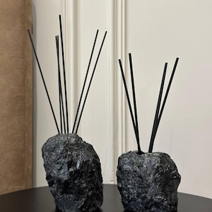 Op de afbeelding: Twee zwarte stenen diffuser bases met zwarte diffuser stokjes. De bases zijn gevormd als rotsen en hebben een ruwe textuur.