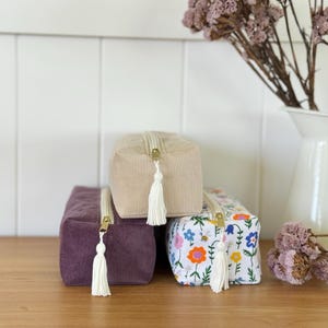May include: Three rectangular cosmetic bags. One is purple corduroy, one is beige corduroy, and the third is white with a floral pattern. Each bag has a gold zipper and a white tassel.