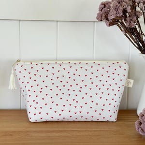 Op de afbeelding: Witte make-uptas met een rood hartjespatroon. De tas heeft een crèmekleurige rits en kwast. Een klein label met het woord "KLEE" is bevestigd. De tas staat op een houten oppervlak.
