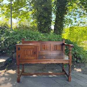 Può includere: Una panca in legno marrone scuro con un piccolo armadietto sul lato sinistro. La panca presenta intagli decorativi e uno schienale con un bracciolo. La panca è all'aperto, con alberi e vegetazione sullo sfondo.