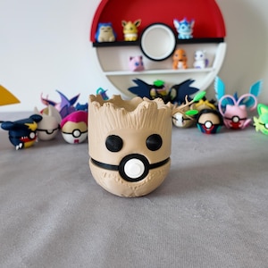 Puede incluir: Una maceta en forma de Groot de color beige con detalles faciales en blanco y negro. La maceta está frente a una estantería temática de Pokémon con varias figuras. La estantería es roja y blanca, con la palabra "Pokemon" en la parte superior.