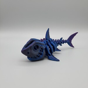 Peut inclure: Figurine de squelette de requin imprimée en 3D, dans des tons de bleu et de violet. La figurine présente des détails squelettiques, des nageoires et un visage souriant. Un objet décoratif unique.