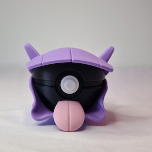 Puede incluir: Una figura de personaje de dibujos animados púrpura y negro con una protuberancia rosa en forma de lengua. La figura tiene un cuerpo esférico con un círculo blanco en el centro y dos cuernos púrpuras.