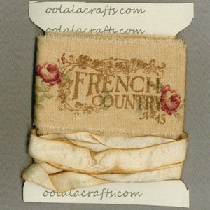 May include: A spool of beige ribbon with a floral print and the text "French Country" in brown lettering. The ribbon is wrapped around a cardboard spool with a white label that says "oolaacrafts.com".