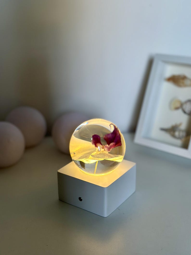 May include: A clear glass sphere with a purple flower inside, resting on a white, illuminated square base. The base has a small, round button on the front. The background includes a white frame with a picture and three round, light-colored objects.