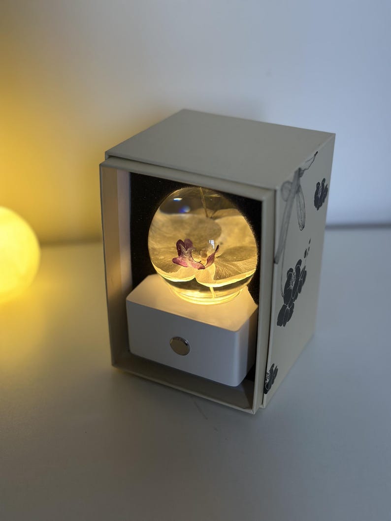 May include: A decorative light featuring a clear glass orb with a purple flower inside, resting on a white base. The orb is illuminated, set within a cream-colored box with floral illustrations. The box is open, revealing the light.
