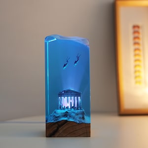 May include: A rectangular lamp featuring a wooden base and a clear blue resin top. Inside the resin, a miniature white temple structure sits on a rocky base, illuminated from below. Two small diver figures are suspended in the blue resin above.