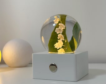 May include: A clear, spherical decorative lamp with a white base. Inside the sphere are white flowers and green leaves. The lamp has a silver button on the front and emits a soft, warm light. The lamp is on a white surface.