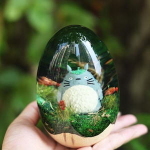 May include: A clear, egg-shaped resin paperweight containing a miniature gray and white Totoro figure. The interior features green moss, small red accents, and a woodland scene encased in transparent material with a smooth, rounded top surface.