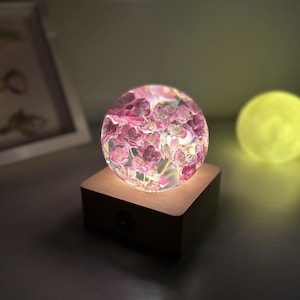 Puede incluir: Una esfera decorativa llena de flores rosas, iluminada desde dentro, descansa sobre una base de madera. La esfera brilla, proyectando una luz suave. Una esfera amarilla está en el fondo. Tema: decoración del hogar.