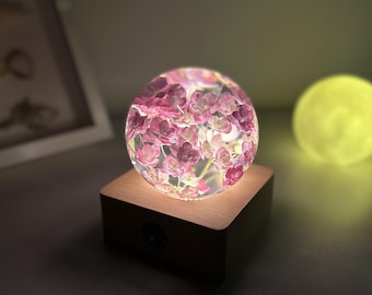 May include: A decorative sphere filled with pink flowers, illuminated from within, rests on a wooden base. The sphere glows, casting a soft light. A yellow sphere is in the background. The overall theme is home decor.