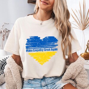 May include: A white t-shirt featuring a blue and yellow heart design. The heart is styled to resemble the Ukrainian flag. The text "Make Empathy Great Again" is printed beneath the heart.