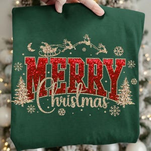 Op de afbeelding: Een donkergroene sweater met de rode glittertekst MERRY en de gouden glittertekst Christmas in script. Het ontwerp bevat een gouden glittersilhouet van de kerstman in een arrenslee, twee gouden glitterkerstbomen en sneeuwvlokken.