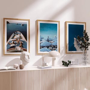 Puede incluir: Tres impresiones artísticas enmarcadas con marcos dorados. Las impresiones representan un yate, sombrillas de playa y un barco en el agua. Las impresiones se exhiben sobre una consola blanca con objetos decorativos.