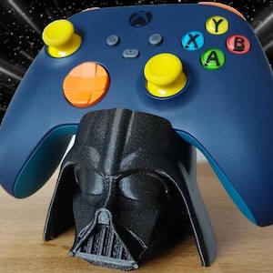 May include: A navy blue Xbox controller with yellow and orange accents rests on a black Darth Vader helmet-shaped stand. The controller has colored buttons and a black background with a starfield.