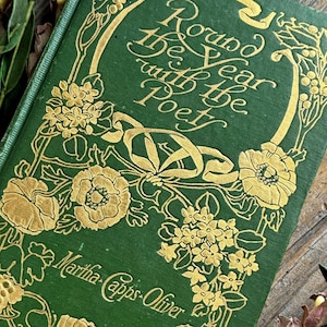 Pode incluir: Um livro verde vintage intitulado "Round the Year with the Poet" com enfeites florais dourados. O nome do autor, Martha Capps-Oliver, tamb&eacute;m est&aacute; impresso na capa. O livro &eacute; cercado por detalhes florais verdes e roxos.