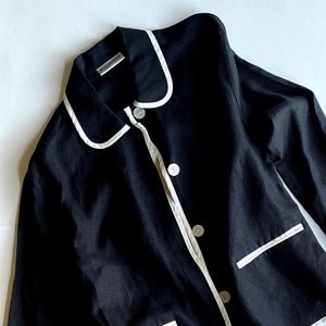 May include: Black blazer with white trim and buttons. The blazer has a classic design with a notched collar and two front pockets. The trim outlines the collar, front, pockets, and cuffs. The fabric appears to be linen.