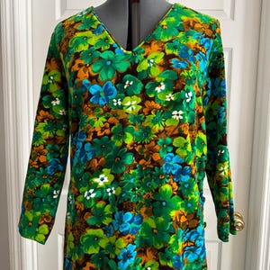 May include: A vintage long-sleeve dress with a V-neck, featuring a vibrant floral pattern. The design includes green, blue, orange, and white flowers on a brown background. The dress is displayed on a mannequin.