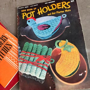 May include: A vintage craft book titled New Book of Pot Holders and Hot Platter Mats, Book No. 274. The black cover features illustrations of three crocheted items: a blue chicken, a green bundle of asparagus, and a yellow squash-shaped pot holder.