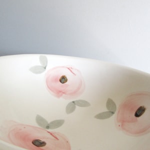 May include: A white ceramic bowl featuring a hand-painted floral pattern. The design consists of soft pink circular flower blossoms with dark brown centers, accented by small, light gray leaves scattered across the interior surface.