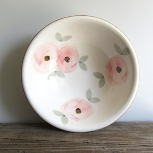 May include: White ceramic bowl with a gold rim, hand-painted with pink floral designs and gray leaves. The bowl has a round shape and is set against a neutral background. A decorative piece for the kitchen or dining room.