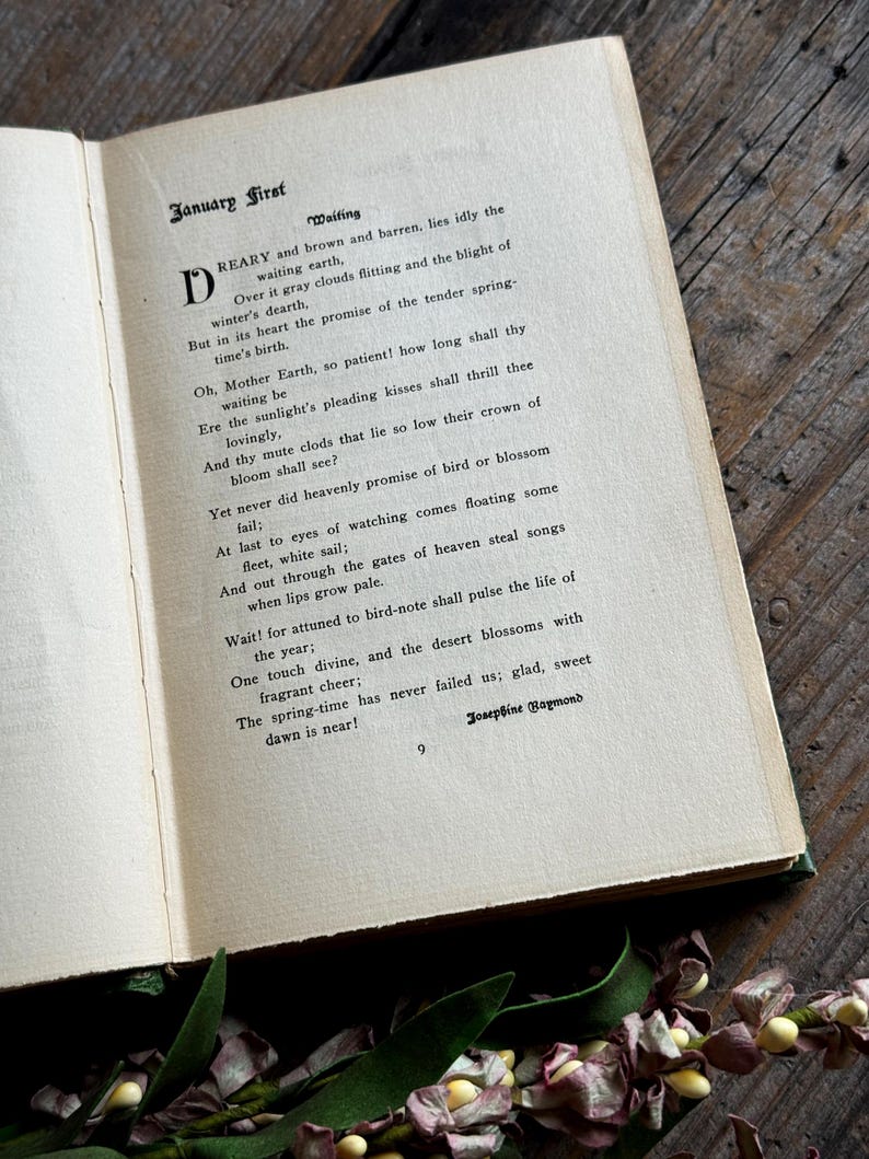 Pode incluir: Um livro aberto com p&aacute;ginas amareladas e envelhecidas, exibindo o poema "January First" de Josephine Raymond. O texto est&aacute; em tinta preta, com uma letra mai&uacute;scula decorativa. O livro repousa sobre uma superf&iacute;cie de madeira escura, com detalhes florais.