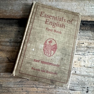 May include: A vintage book titled "Essentials of English First Book" with red lettering and a circular illustration. The book has a tan linen cover and is set against a weathered wooden surface, evoking a sense of history and education.