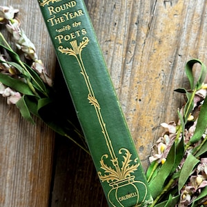 Pode incluir: Um livro verde vintage intitulado "Round the Year with the Poets" com letras douradas e detalhes decorativos. O livro est&aacute; sobre uma superf&iacute;cie de madeira, com detalhes florais no fundo.