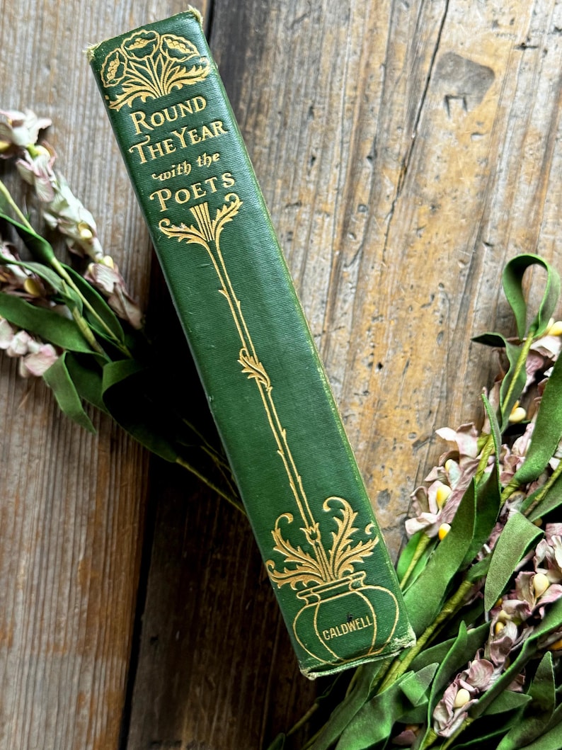 Pode incluir: Um livro verde vintage intitulado "Round the Year with the Poets" com letras douradas e detalhes decorativos. O livro est&aacute; sobre uma superf&iacute;cie de madeira, com detalhes florais no fundo.