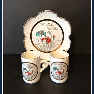 May include: A white ceramic plate with a gold rim and a floral design. The plate has the text "To Mother With Love" on it. Two white ceramic mugs with gold rims and the same floral design and text are also shown.