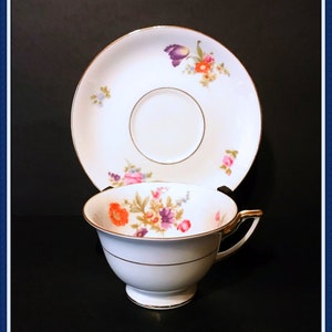 May include: A white teacup and saucer with a floral pattern in shades of purple, orange, and yellow. The cup and saucer have a gold trim.